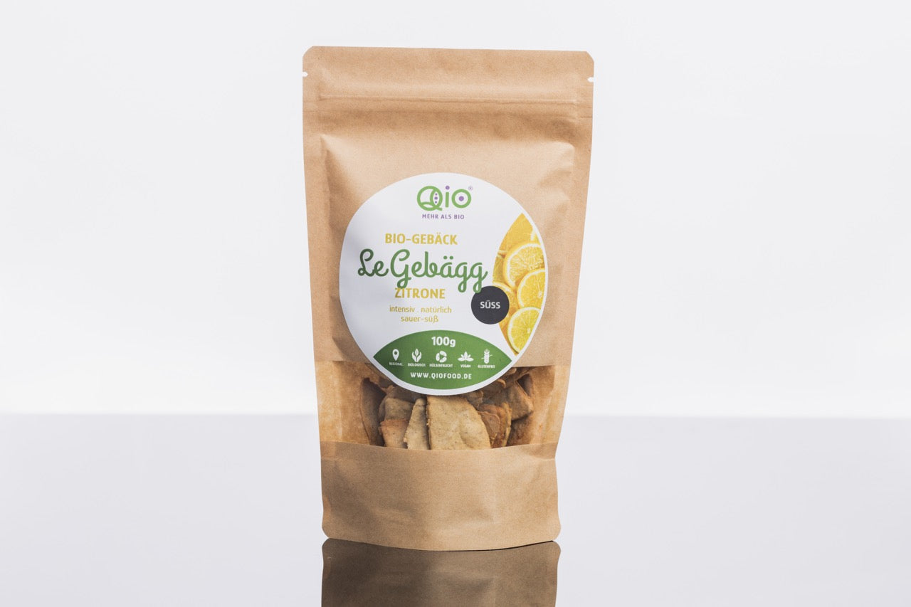 Eine braune Papiertüte mit der Aufschrift „LeGebäck – Zitrone 100 g“ und einem grün-weiß-gelben Etikett enthält vegane Bio-Zitronenkekse von Qio Food, präsentiert auf einem Regal.
