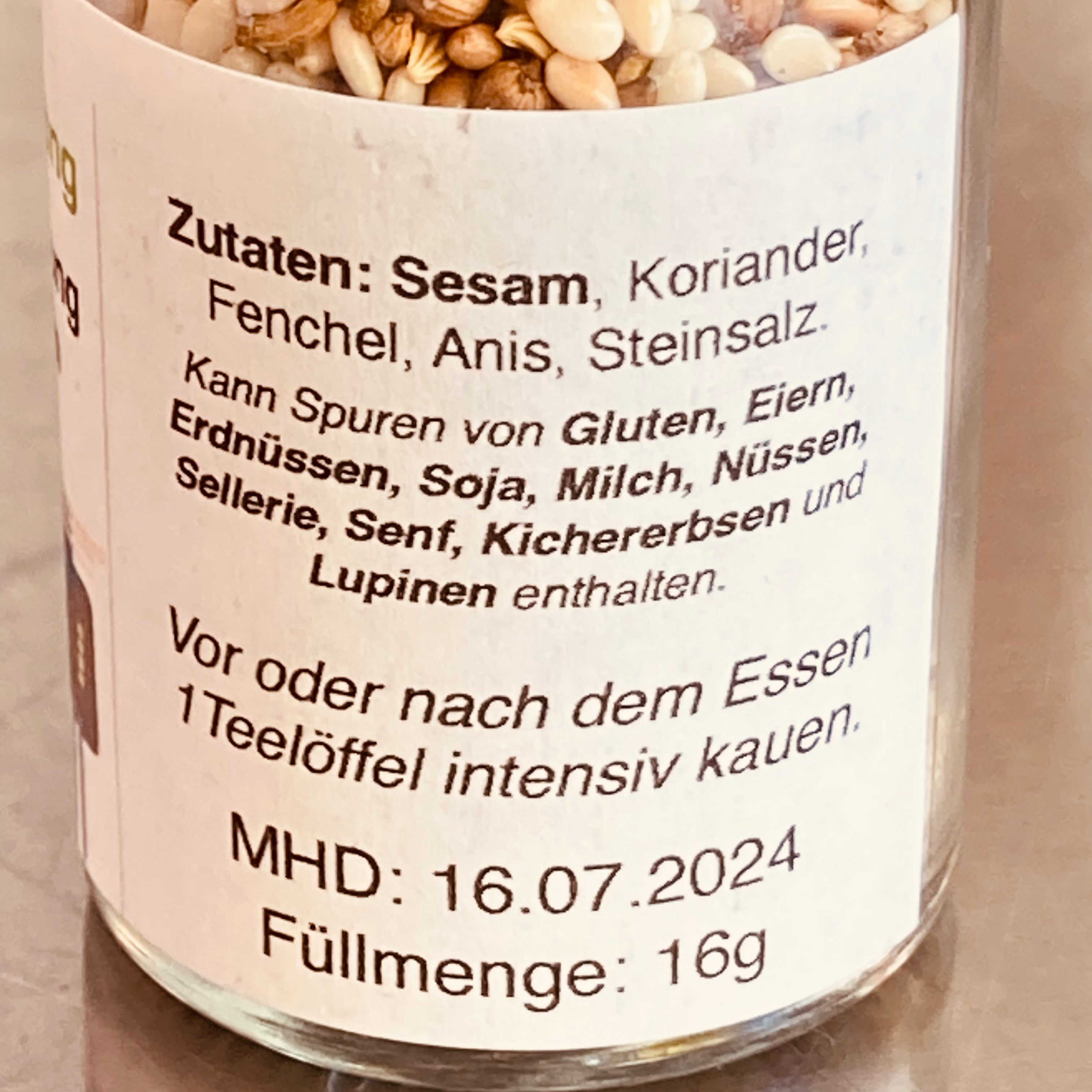 Nahaufnahme des Etiketts eines Qio Food-Glases mit einer Zutatenliste auf Deutsch, beispielsweise Sesam, Koriander und verschiedene Allergene, wobei das Mindesthaltbarkeitsdatum 16.07.2024 sichtbar ist.