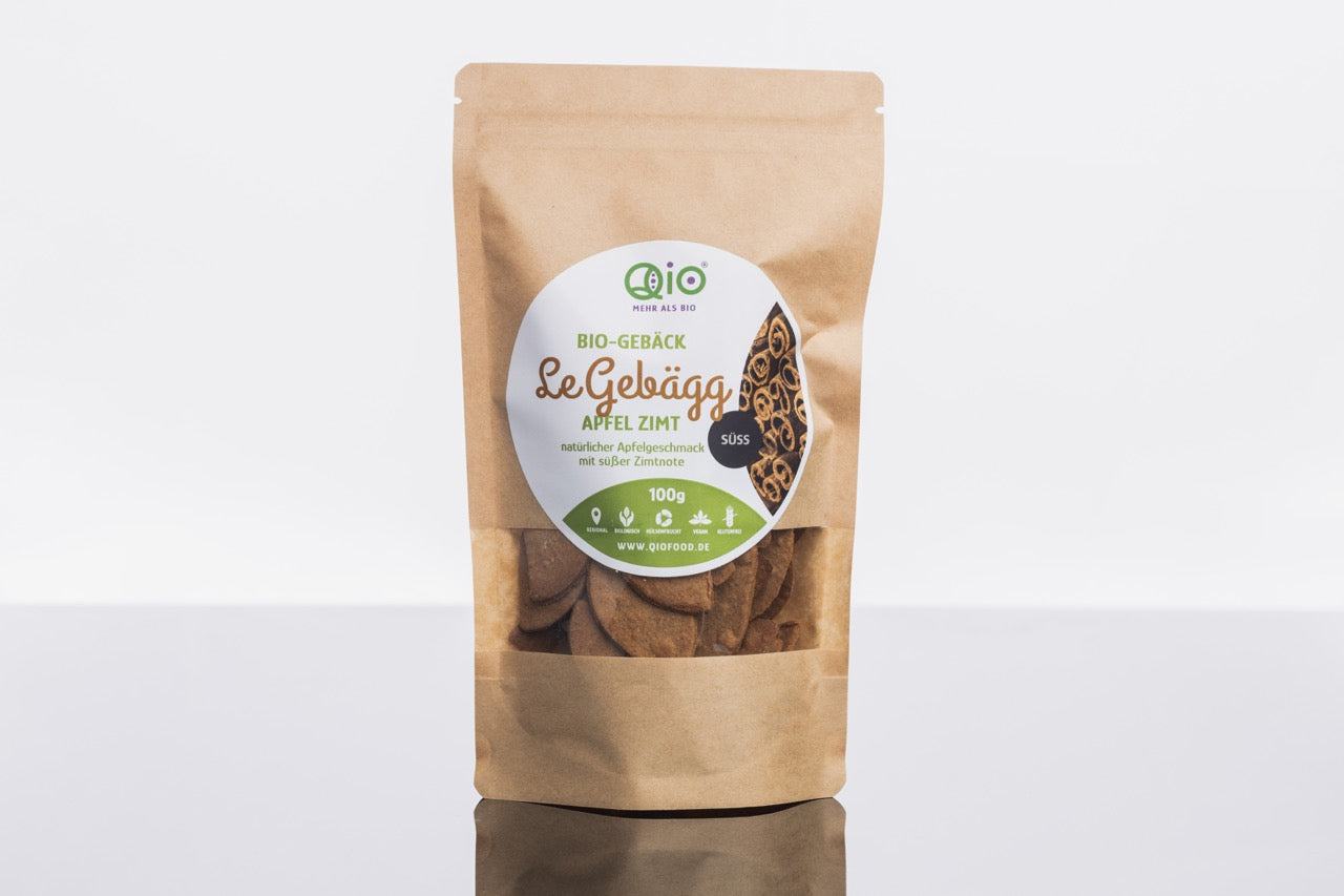 Eine braune Papiertüte von Qio Food mit einem Etikett für „LeGebägg - Apfel Zimt 100g“ mit Keksen, grünem Text und einem Logo oben drauf. Die Tüte ist nachhaltig.